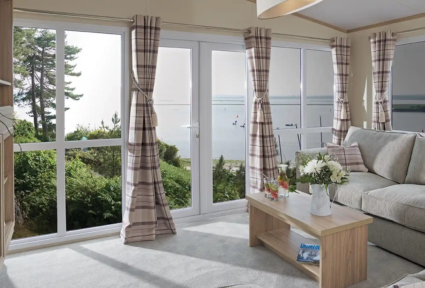 The image shows a cosy, modern living room with a soft, neutral color scheme. The room has large windows, providing a beautiful view of the outdoors, possibly a lake or river, with sailboats visible in the distance. The furniture includes a comfortable sofa with cushions and a simple wooden coffee table, adorned with a flower arrangement and drinks. The curtains are checked in a subtle, pastel color, and the light from the large window creates a bright and airy atmosphere. The space feels inviting, tranquil, and connected to nature outside.