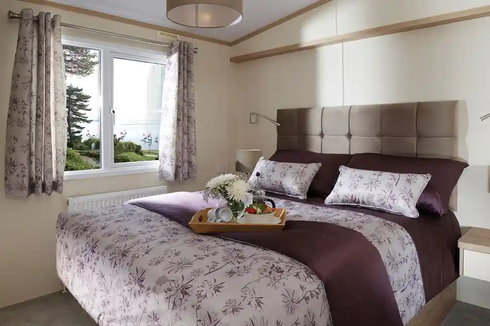 This image presents a serene and cozy master bedroom. The bed is beautifully made with a rich purple and floral-patterned duvet, complemented by matching pillows. The headboard is upholstered in a soft beige fabric, adding a touch of luxury to the room. The space is brightened by natural light streaming in through a large window, offering a tranquil view of the outdoors with trees and water visible. A small tray with a cup of tea and some fruit adds a welcoming and homely touch, making the room feel like the perfect retreat. The overall atmosphere is peaceful and elegantly designed.