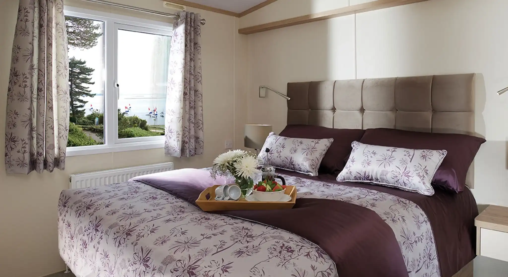 This image presents a serene and cozy master bedroom. The bed is beautifully made with a rich purple and floral-patterned duvet, complemented by matching pillows. The headboard is upholstered in a soft beige fabric, adding a touch of luxury to the room. The space is brightened by natural light streaming in through a large window, offering a tranquil view of the outdoors with trees and water visible. A small tray with a cup of tea and some fruit adds a welcoming and homely touch, making the room feel like the perfect retreat. The overall atmosphere is peaceful and elegantly designed.