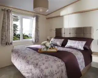 This image presents a serene and cozy master bedroom. The bed is beautifully made with a rich purple and floral-patterned duvet, complemented by matching pillows. The headboard is upholstered in a soft beige fabric, adding a touch of luxury to the room. The space is brightened by natural light streaming in through a large window, offering a tranquil view of the outdoors with trees and water visible. A small tray with a cup of tea and some fruit adds a welcoming and homely touch, making the room feel like the perfect retreat. The overall atmosphere is peaceful and elegantly designed.