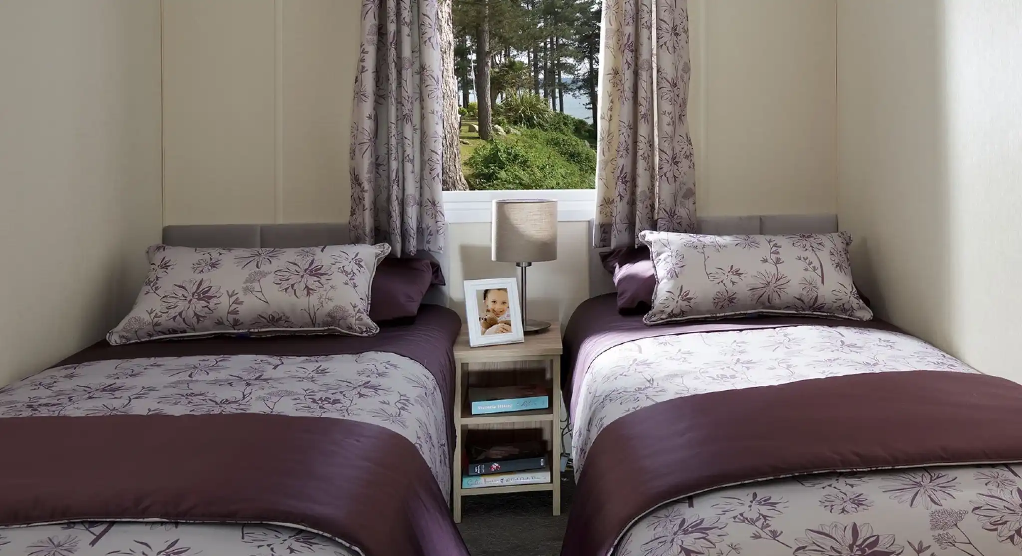 This image features a charming twin bedroom with two single beds, each dressed in elegant purple and floral-patterned bedding. The soft color palette, with shades of purple and lavender, creates a calming atmosphere. Each bed has a matching pillow and a small side table with a lamp and a framed photo, adding a personal touch. The room is well-lit by natural light pouring in through the window, which offers a view of trees outside. The simple yet functional layout makes this space cozy and inviting, perfect for guests or children.