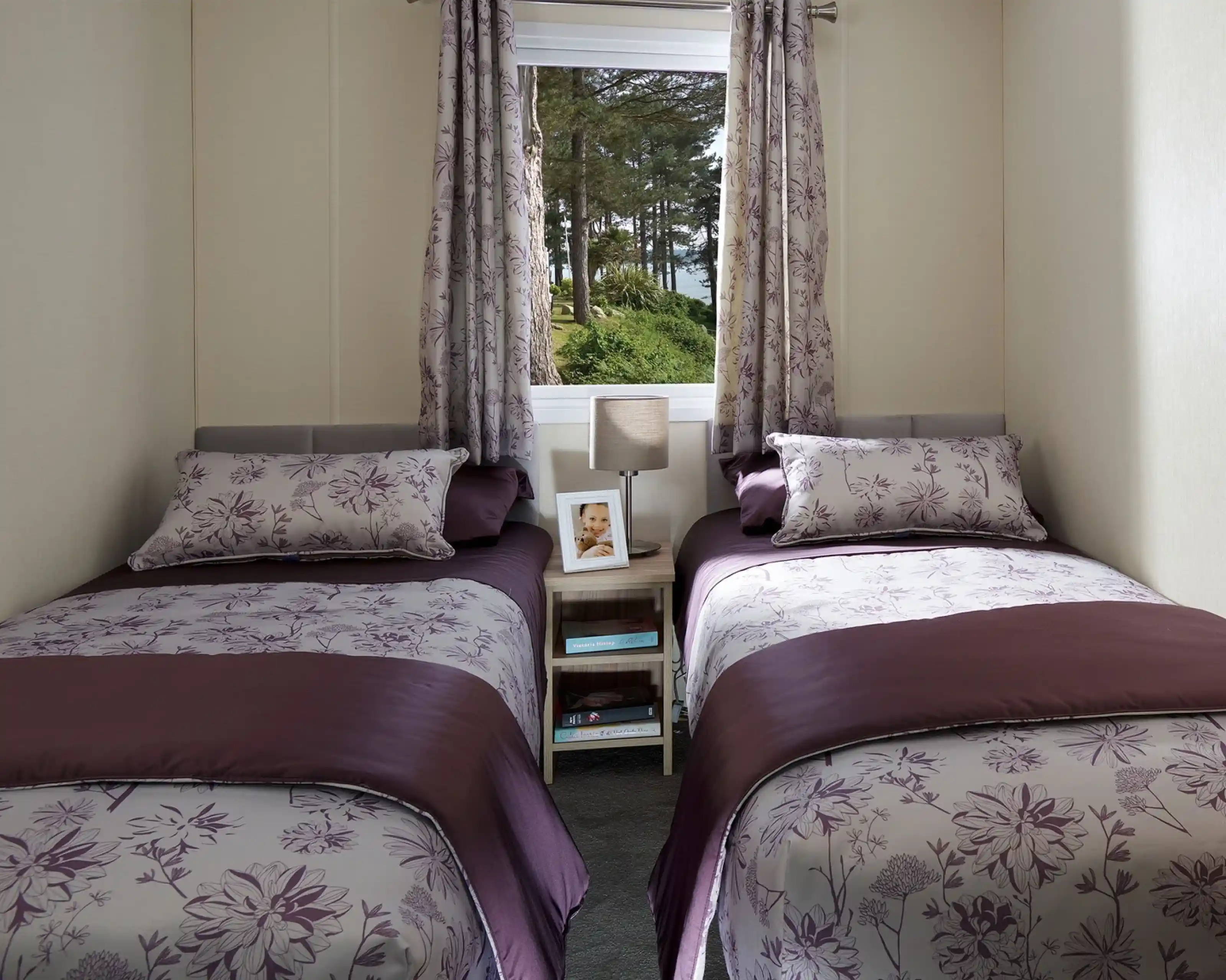 This image features a charming twin bedroom with two single beds, each dressed in elegant purple and floral-patterned bedding. The soft color palette, with shades of purple and lavender, creates a calming atmosphere. Each bed has a matching pillow and a small side table with a lamp and a framed photo, adding a personal touch. The room is well-lit by natural light pouring in through the window, which offers a view of trees outside. The simple yet functional layout makes this space cozy and inviting, perfect for guests or children.
