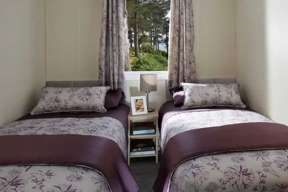 This image features a charming twin bedroom with two single beds, each dressed in elegant purple and floral-patterned bedding. The soft color palette, with shades of purple and lavender, creates a calming atmosphere. Each bed has a matching pillow and a small side table with a lamp and a framed photo, adding a personal touch. The room is well-lit by natural light pouring in through the window, which offers a view of trees outside. The simple yet functional layout makes this space cozy and inviting, perfect for guests or children.
