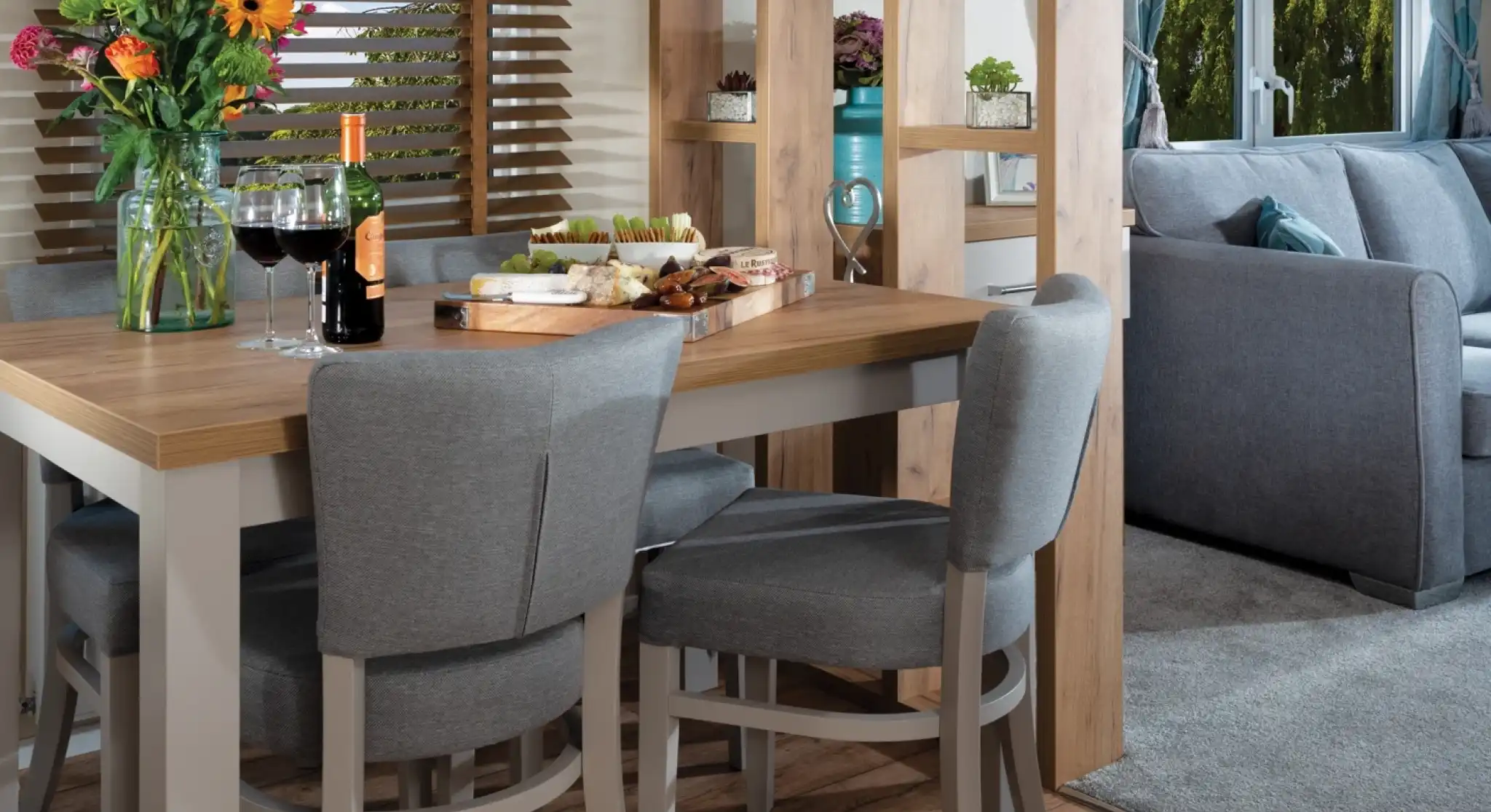 Modern dining area with a wooden table, gray chairs, and colorful flowers. Cheese and wine set a cozy, inviting mood. Bright window view outside.