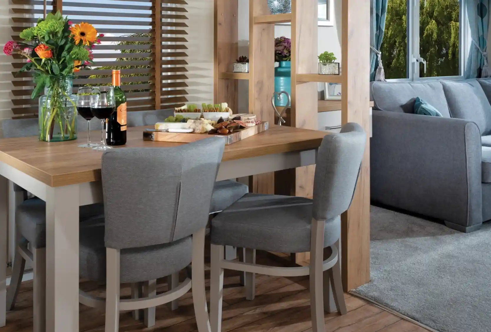 Modern dining area with a wooden table, gray chairs, and colorful flowers. Cheese and wine set a cozy, inviting mood. Bright window view outside.