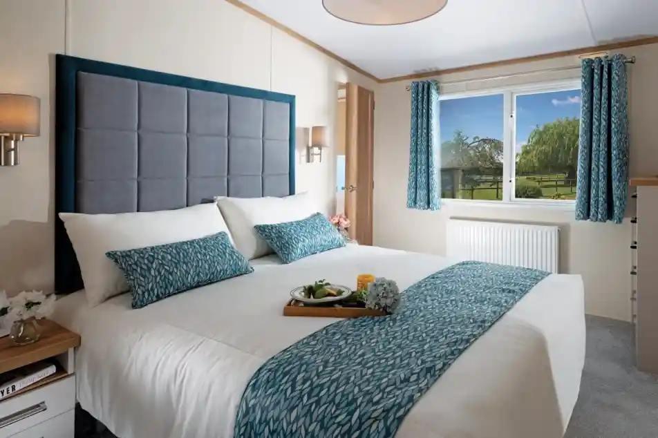 Bright bedroom with a large, neatly made bed featuring blue-patterned accents. A window with matching curtains reveals a scenic garden view. Cozy and inviting atmosphere.
