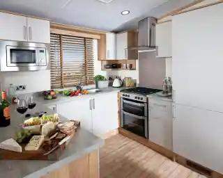 Modern kitchen with white cabinets, wooden accents, and grey countertops. A platter with assorted cheeses, grapes, and two glasses of red wine sits on the counter. A window with wooden blinds lets light in, creating a warm, inviting atmosphere.