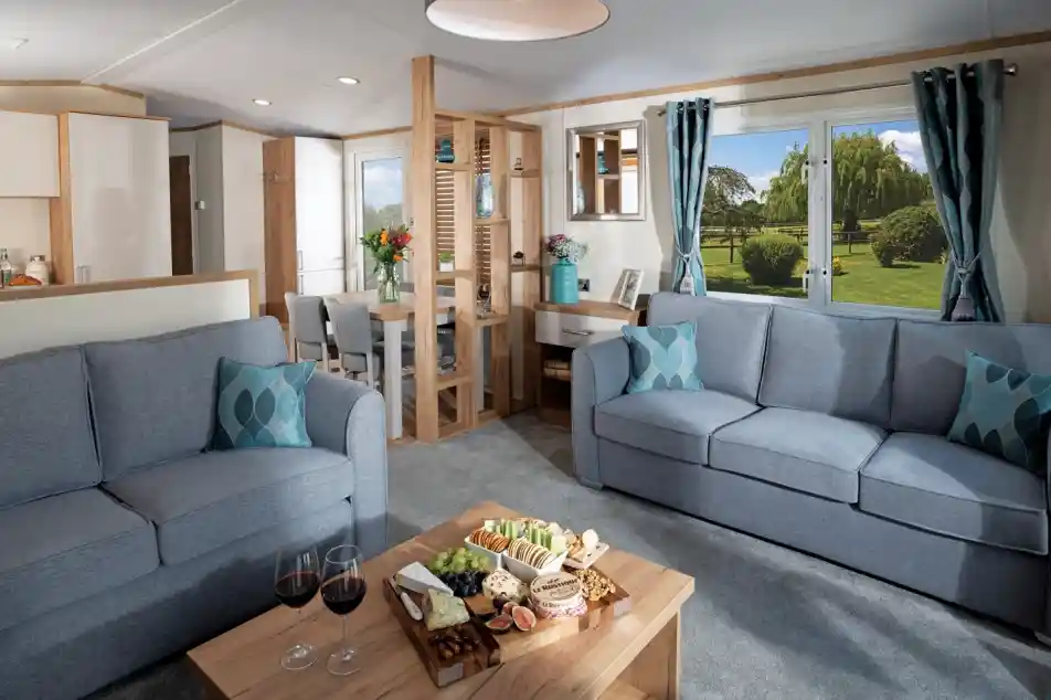 Modern living room with two blue sofas, wood coffee table with snacks and wine; large window showing a lush garden. Bright and inviting atmosphere.