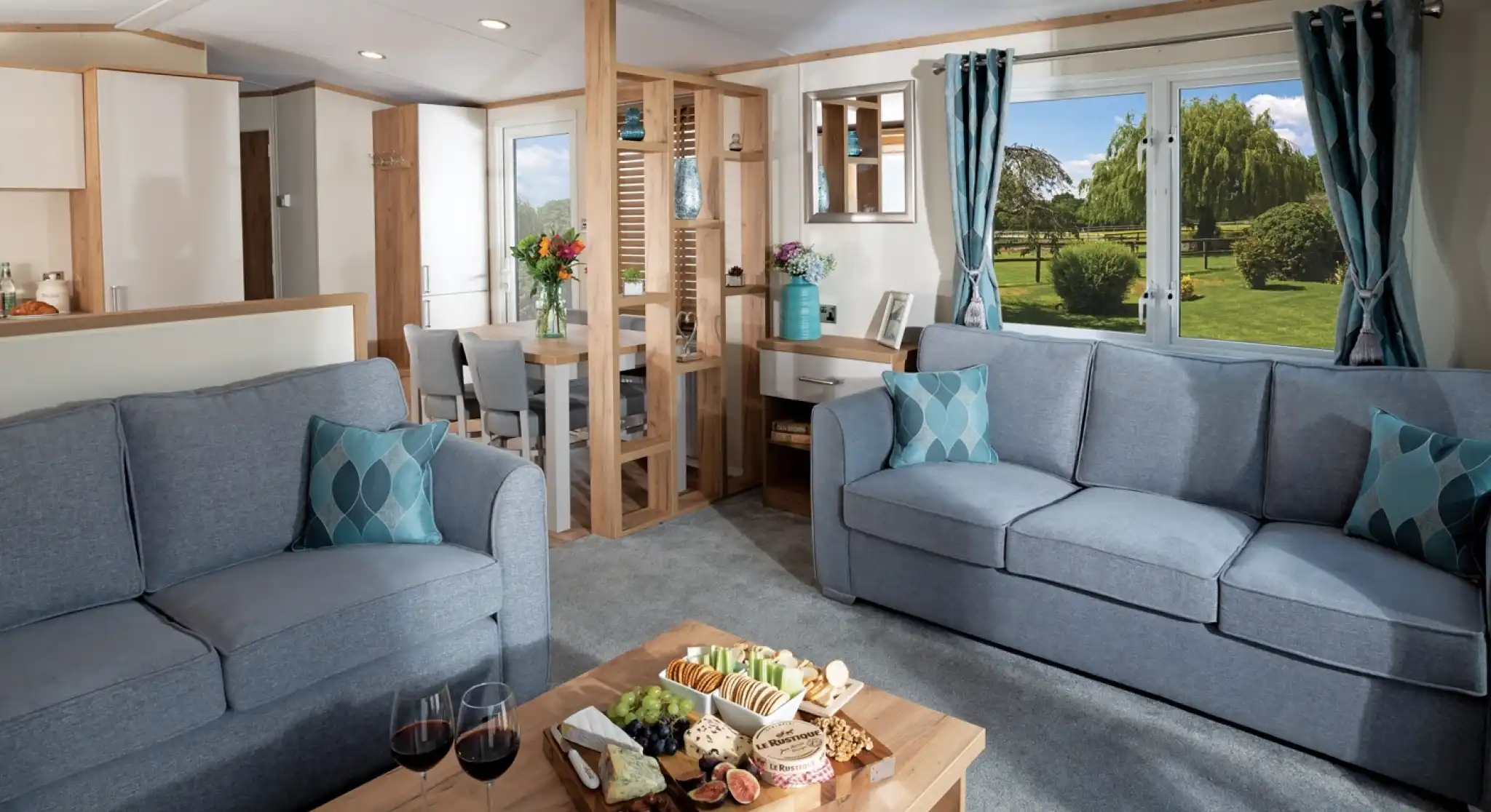 Modern living room with two blue sofas, wood coffee table with snacks and wine; large window showing a lush garden. Bright and inviting atmosphere.