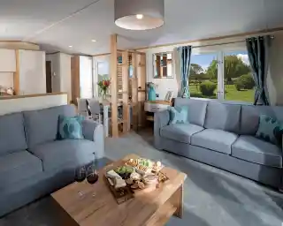 Modern living room with two blue sofas, wood coffee table with snacks and wine; large window showing a lush garden. Bright and inviting atmosphere.