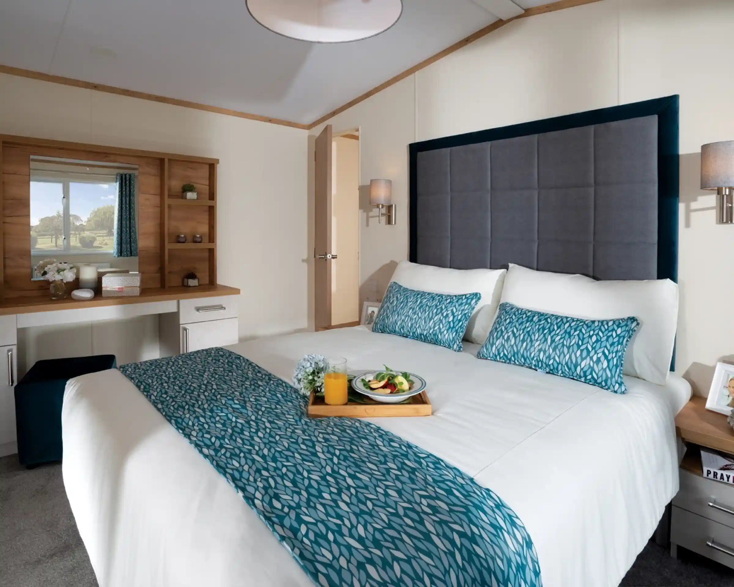 Modern bedroom with a large bed featuring blue patterned accents. A tray with breakfast sits on the bed. A vanity and scenic window view create a serene ambiance.