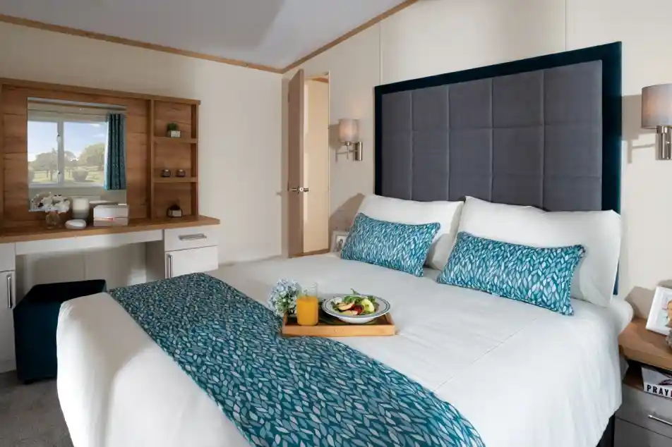 Modern bedroom with a large bed featuring blue patterned accents. A tray with breakfast sits on the bed. A vanity and scenic window view create a serene ambiance.