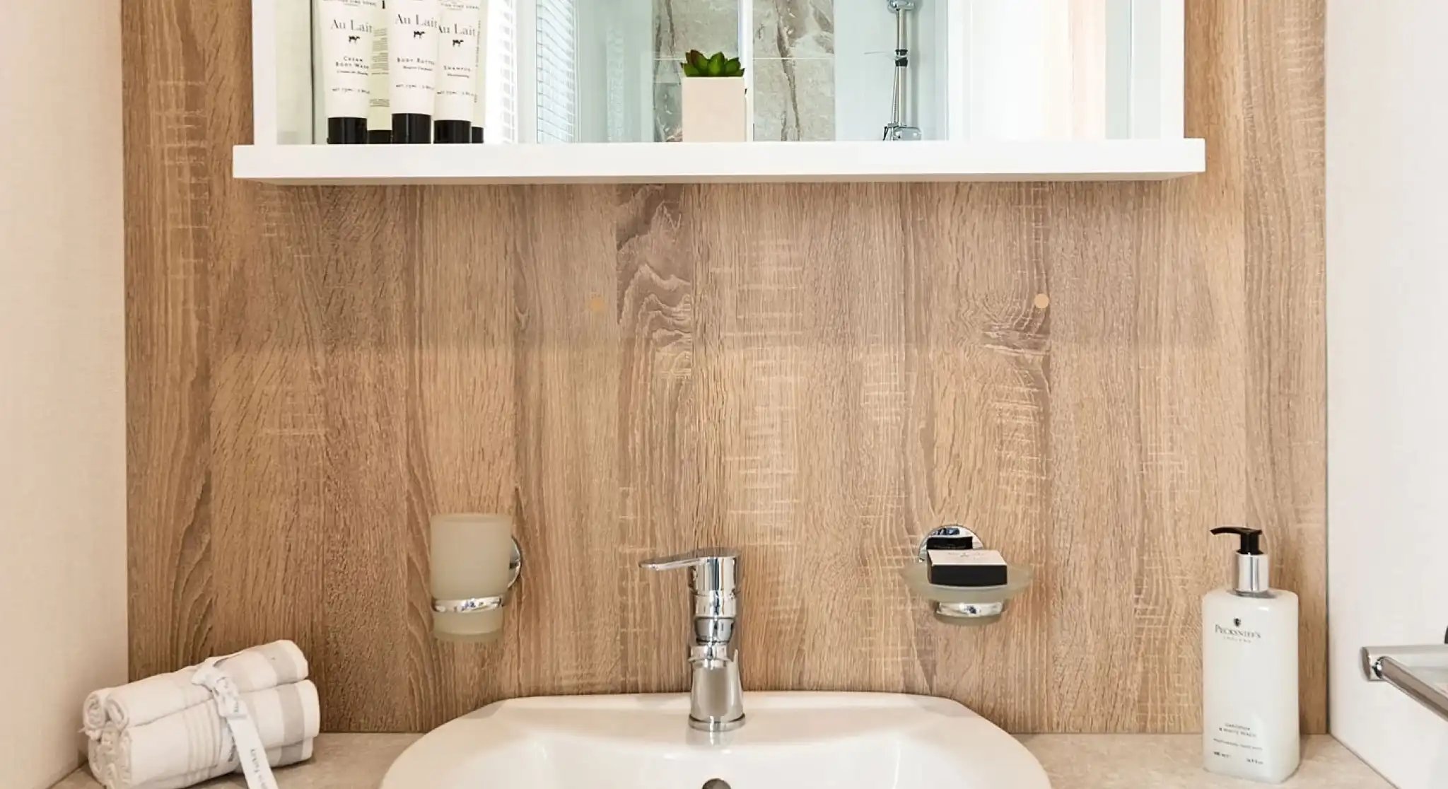 Modern bathroom sink area with a chrome tap, mounted soap dispenser, and beige countertop. A wooden-style wall panel holds a shelf with toiletries and a small potted plant. A shower enclosure with glass doors is reflected in the mirror above.