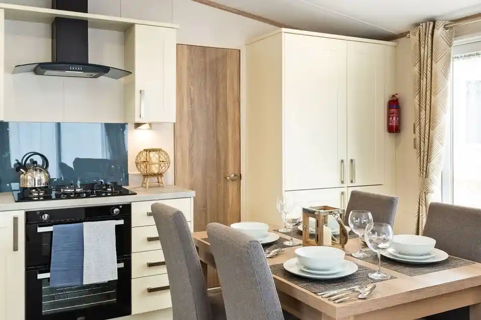 Modern kitchen and dining area with cream cabinets, built-in oven, gas stove, kettle, and a wooden dining table set for four with grey upholstered chairs and place settings.