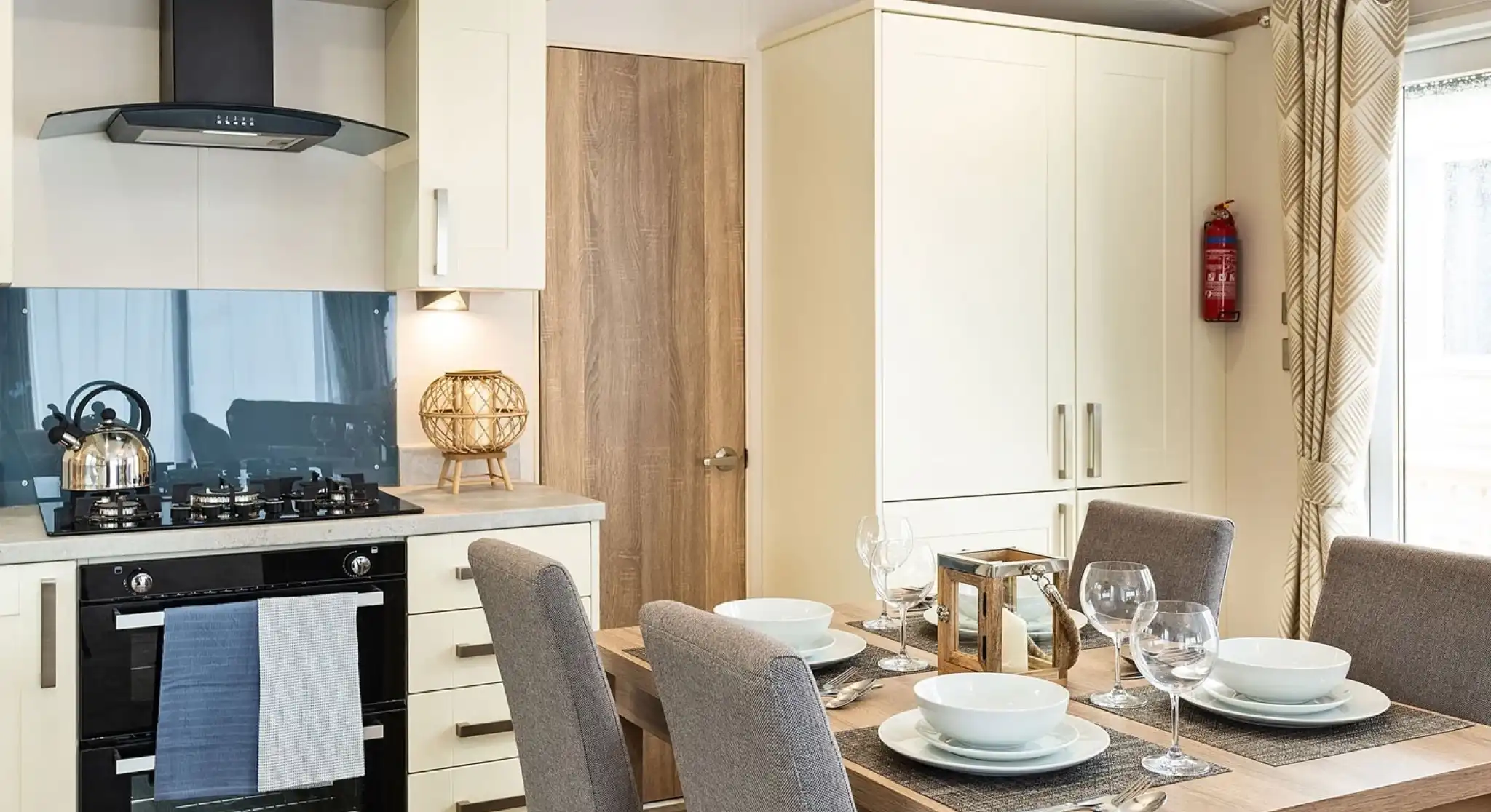 Modern kitchen and dining area with cream cabinets, built-in oven, gas stove, kettle, and a wooden dining table set for four with grey upholstered chairs and place settings.