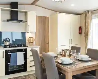 Modern kitchen and dining area with cream cabinets, built-in oven, gas stove, kettle, and a wooden dining table set for four with grey upholstered chairs and place settings.