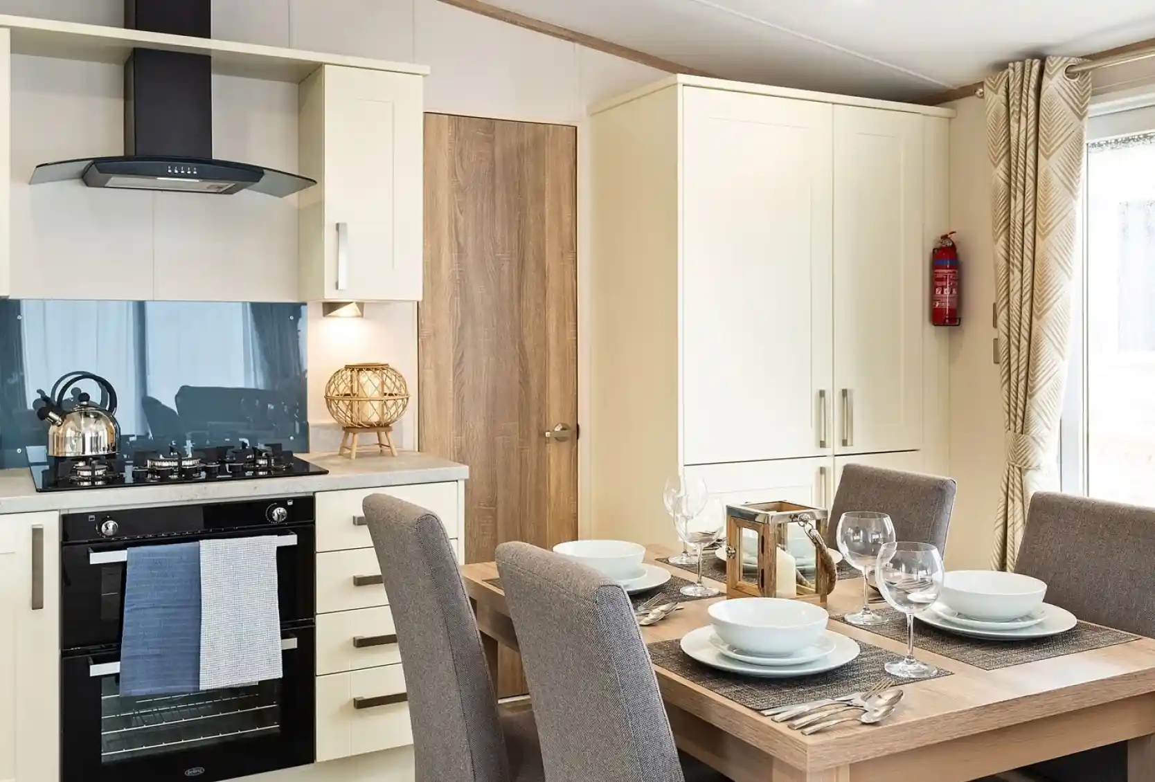 Modern kitchen and dining area with cream cabinets, built-in oven, gas stove, kettle, and a wooden dining table set for four with grey upholstered chairs and place settings.