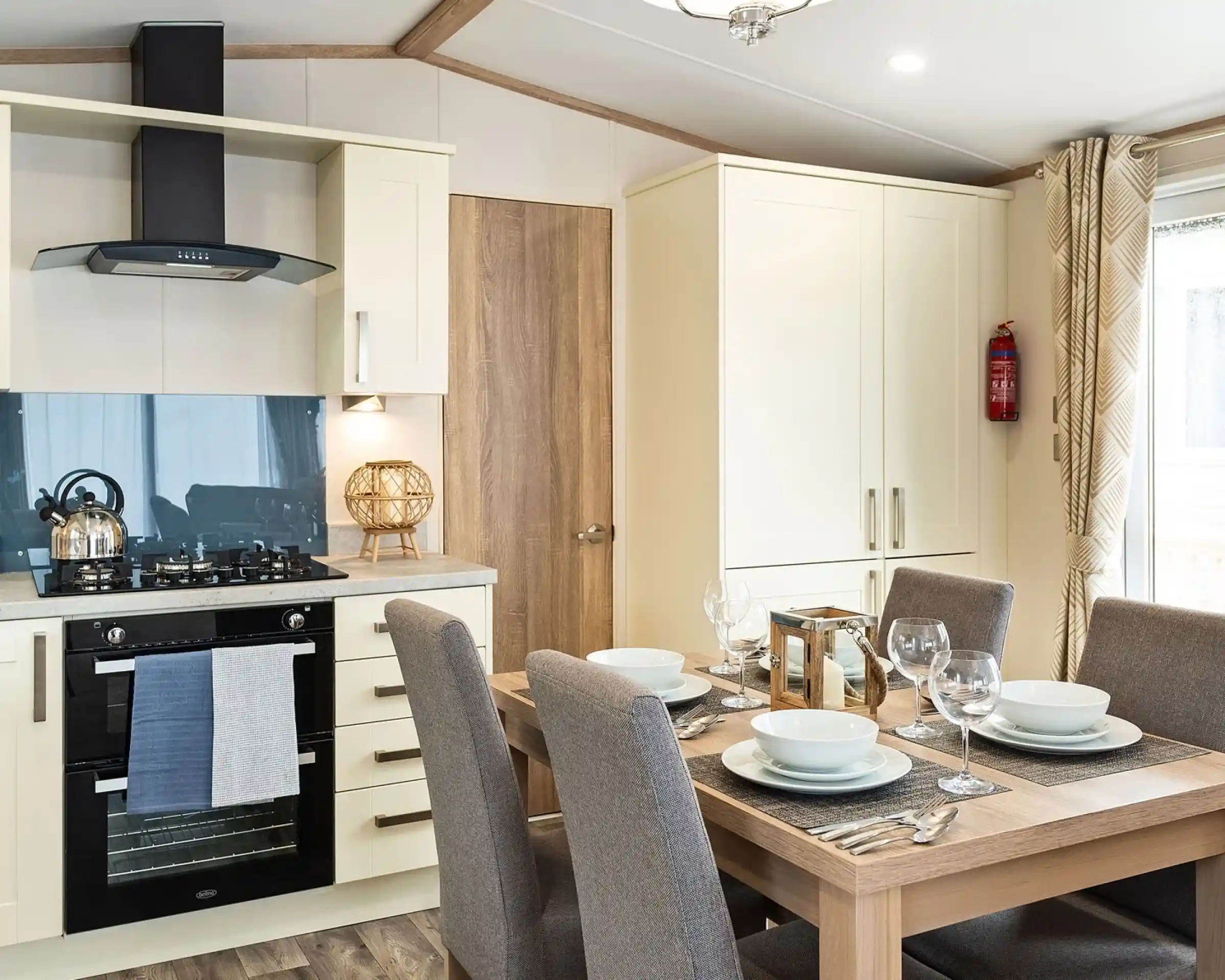 Modern kitchen and dining area with cream cabinets, built-in oven, gas stove, kettle, and a wooden dining table set for four with grey upholstered chairs and place settings.