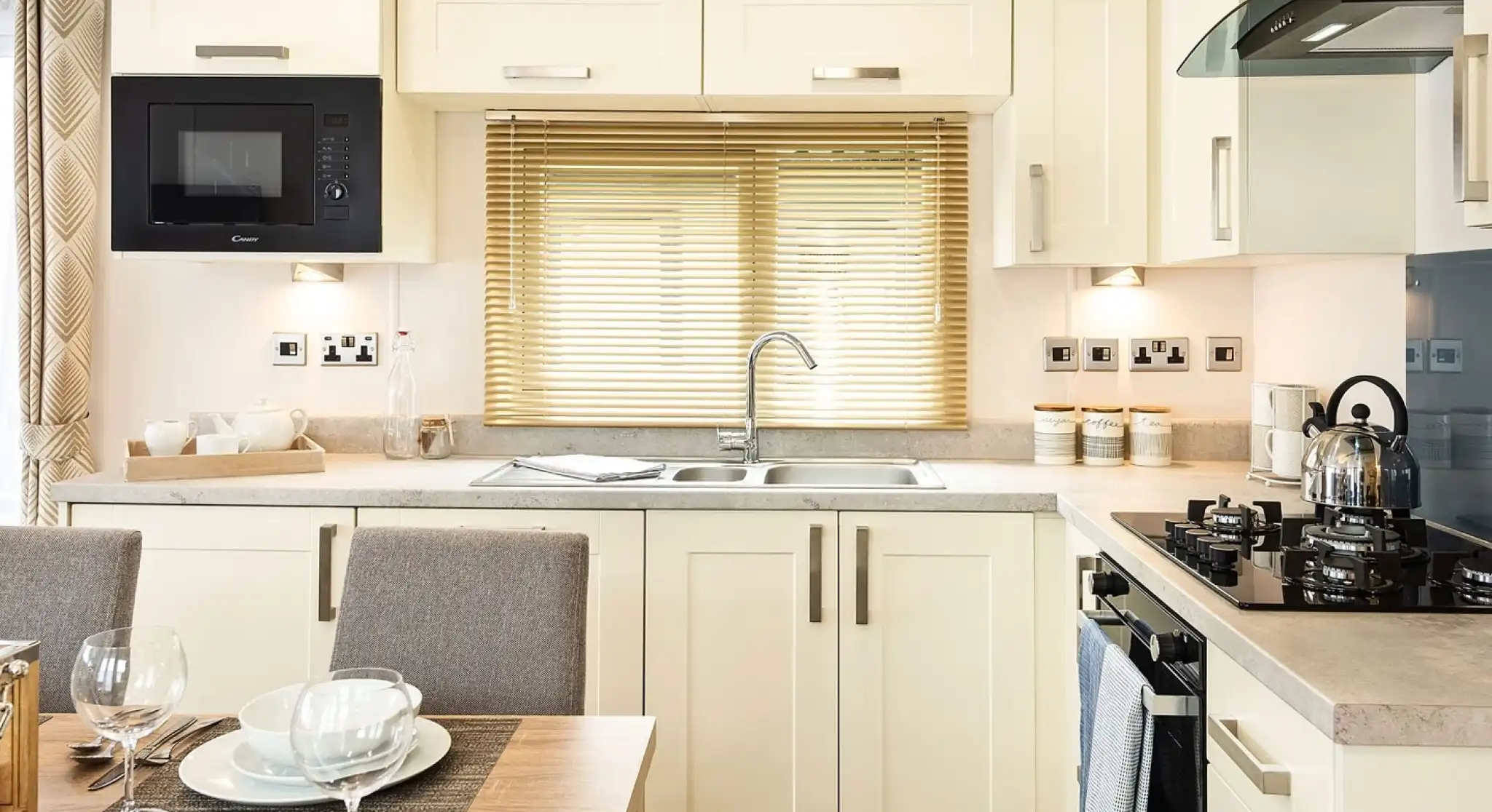Bright modern kitchen with cream cabinets, built-in microwave, stainless steel sink under a window with blinds, gas stove with kettle, and a dining table set with bowls, plates, and wine glasses.