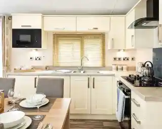 Bright modern kitchen with cream cabinets, built-in microwave, stainless steel sink under a window with blinds, gas stove with kettle, and a dining table set with bowls, plates, and wine glasses.