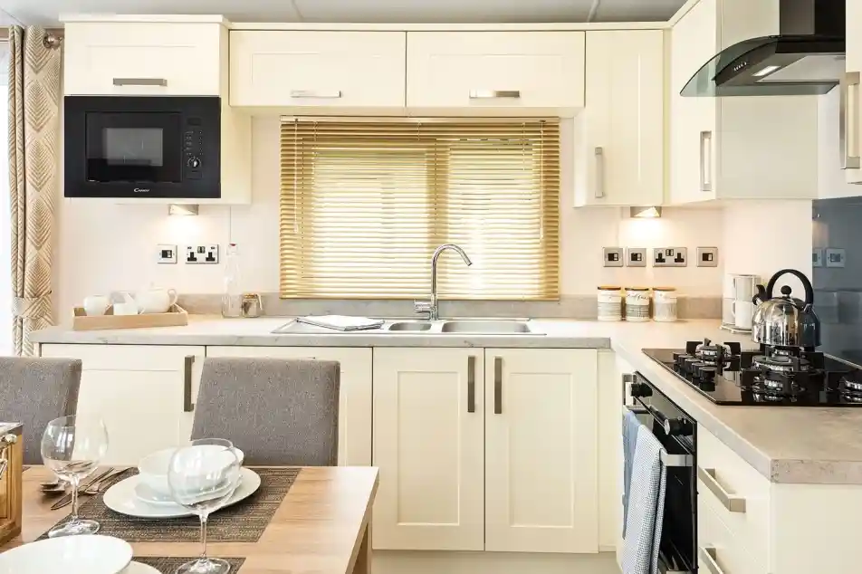 Bright modern kitchen with cream cabinets, built-in microwave, stainless steel sink under a window with blinds, gas stove with kettle, and a dining table set with bowls, plates, and wine glasses.