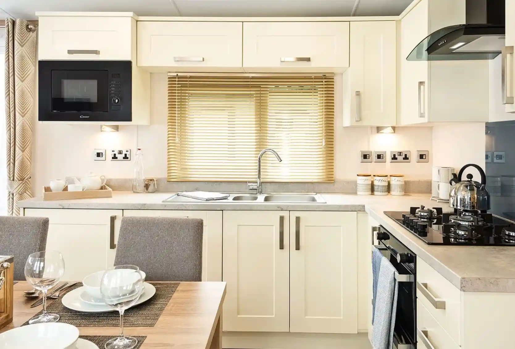 Bright modern kitchen with cream cabinets, built-in microwave, stainless steel sink under a window with blinds, gas stove with kettle, and a dining table set with bowls, plates, and wine glasses.