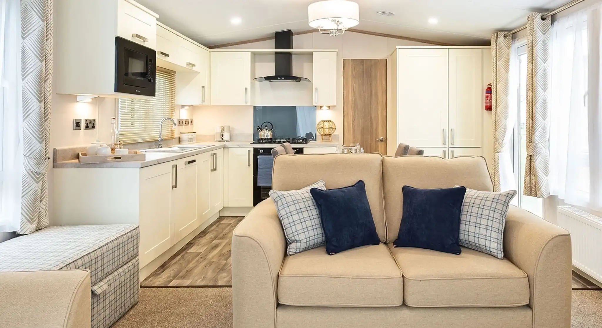Modern open-plan living space inside a mobile home or caravan, featuring a beige two-seater sofa with navy and plaid cushions in the foreground. The kitchen area in the background includes cream cabinets, a built-in microwave, stove with hood, and a small dining area. Large windows with patterned curtains provide ample natural light, and the room is decorated in neutral tones with contemporary furnishings.