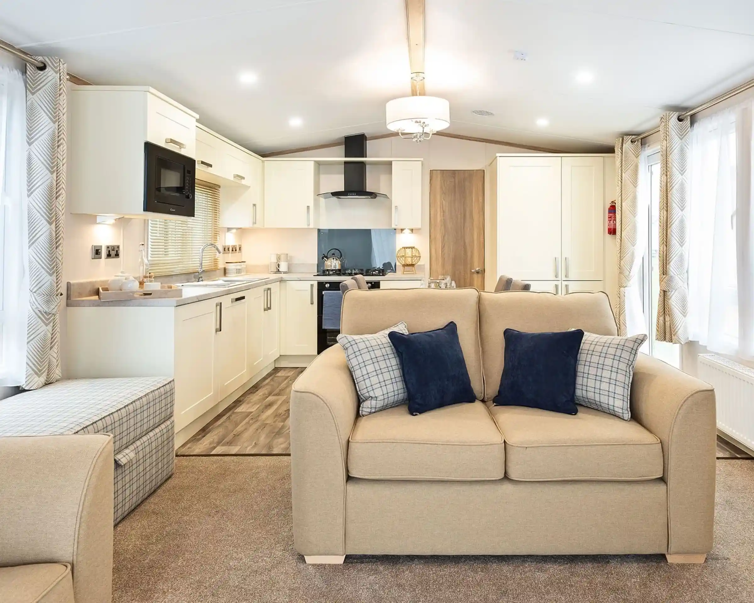Modern open-plan living space inside a mobile home or caravan, featuring a beige two-seater sofa with navy and plaid cushions in the foreground. The kitchen area in the background includes cream cabinets, a built-in microwave, stove with hood, and a small dining area. Large windows with patterned curtains provide ample natural light, and the room is decorated in neutral tones with contemporary furnishings.