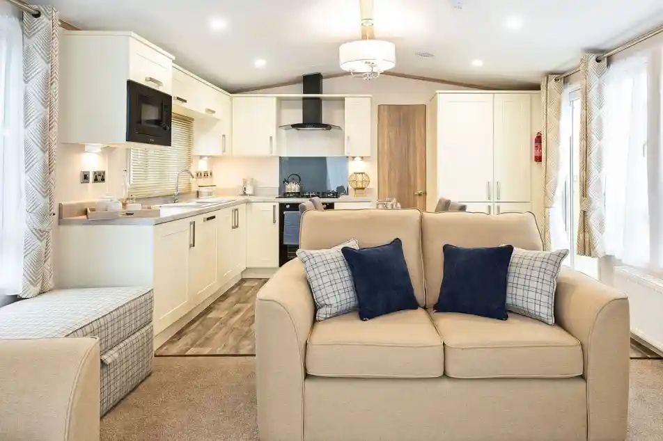 Modern open-plan living space inside a mobile home or caravan, featuring a beige two-seater sofa with navy and plaid cushions in the foreground. The kitchen area in the background includes cream cabinets, a built-in microwave, stove with hood, and a small dining area. Large windows with patterned curtains provide ample natural light, and the room is decorated in neutral tones with contemporary furnishings.