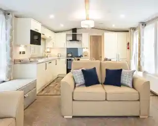 Modern open-plan living space inside a mobile home or caravan, featuring a beige two-seater sofa with navy and plaid cushions in the foreground. The kitchen area in the background includes cream cabinets, a built-in microwave, stove with hood, and a small dining area. Large windows with patterned curtains provide ample natural light, and the room is decorated in neutral tones with contemporary furnishings.