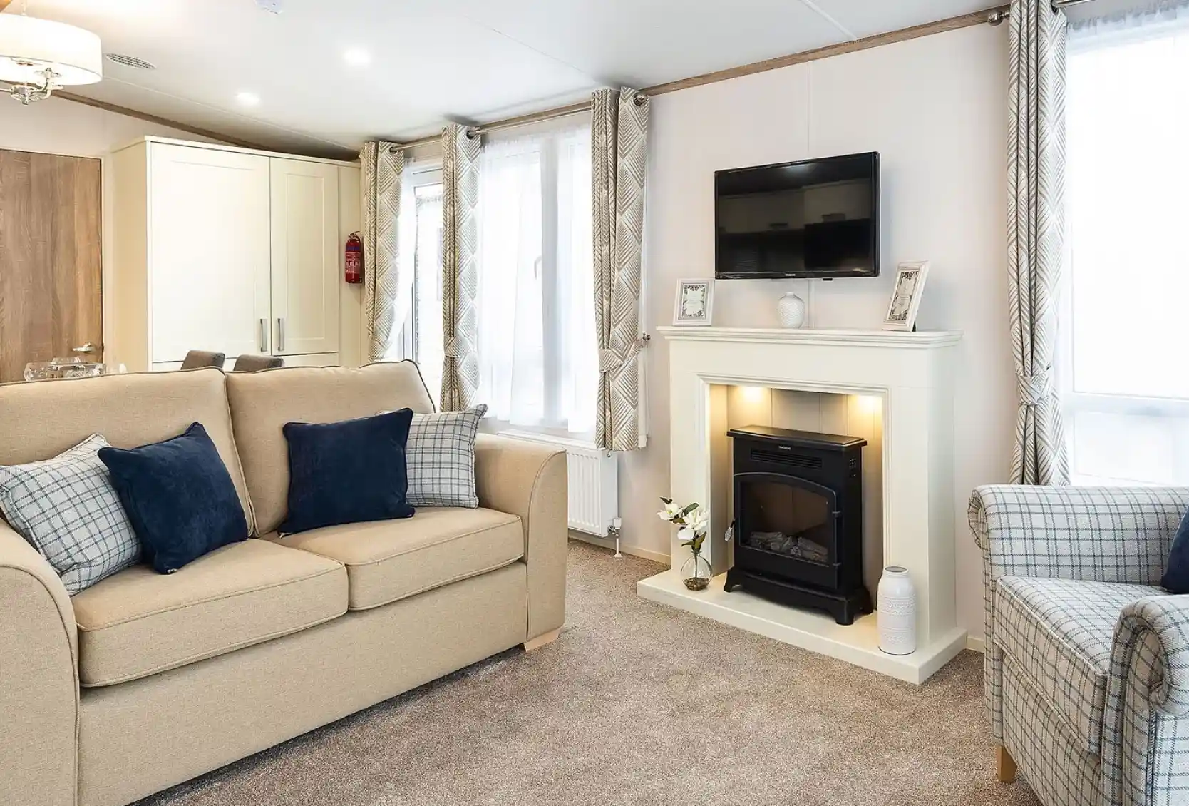 Cozy living room with beige sofa, armchair, and navy and plaid cushions, featuring a white fireplace with built-in electric fire, wall-mounted TV above, and large windows with patterned curtains.