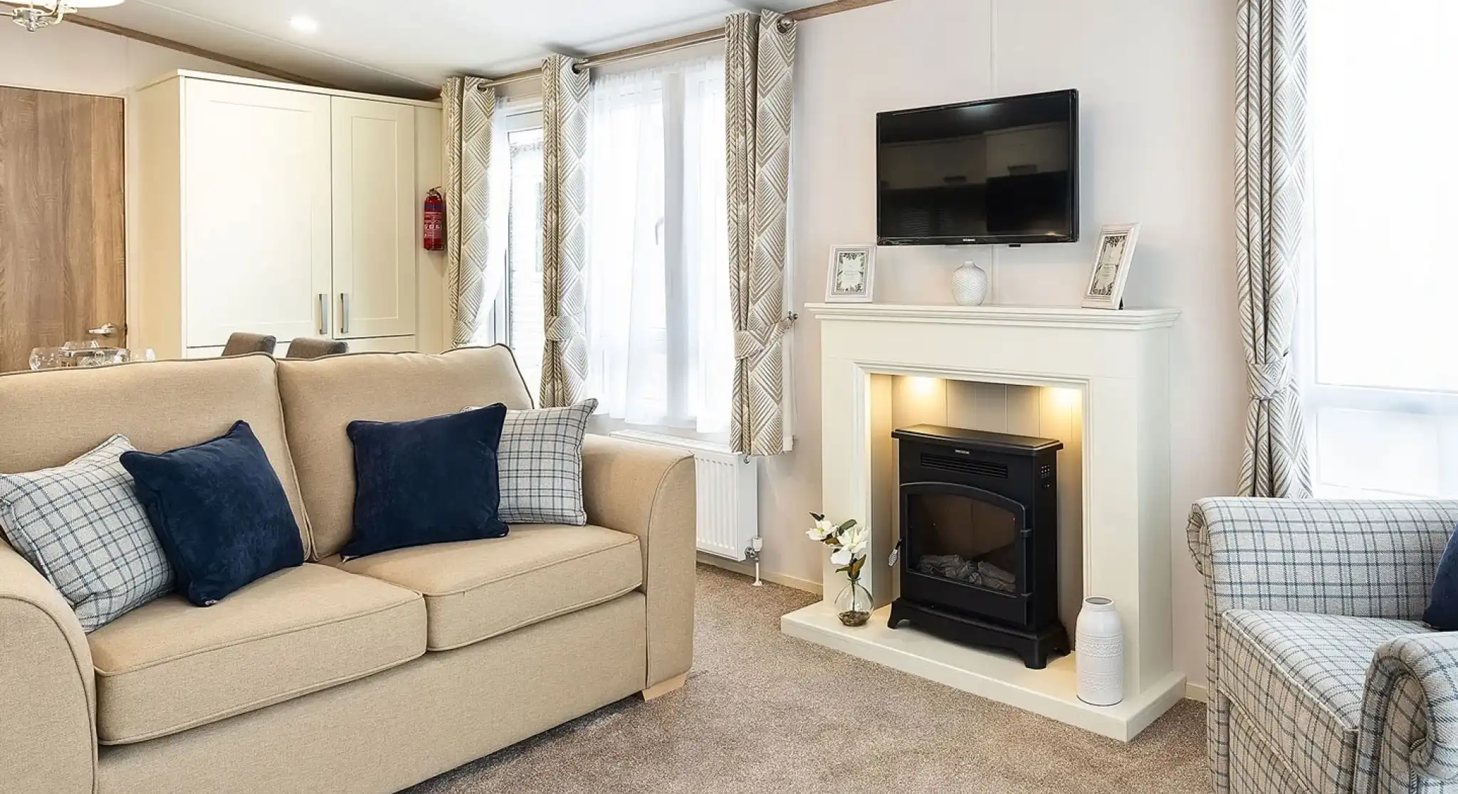 Cozy living room with beige sofa, armchair, and navy and plaid cushions, featuring a white fireplace with built-in electric fire, wall-mounted TV above, and large windows with patterned curtains.