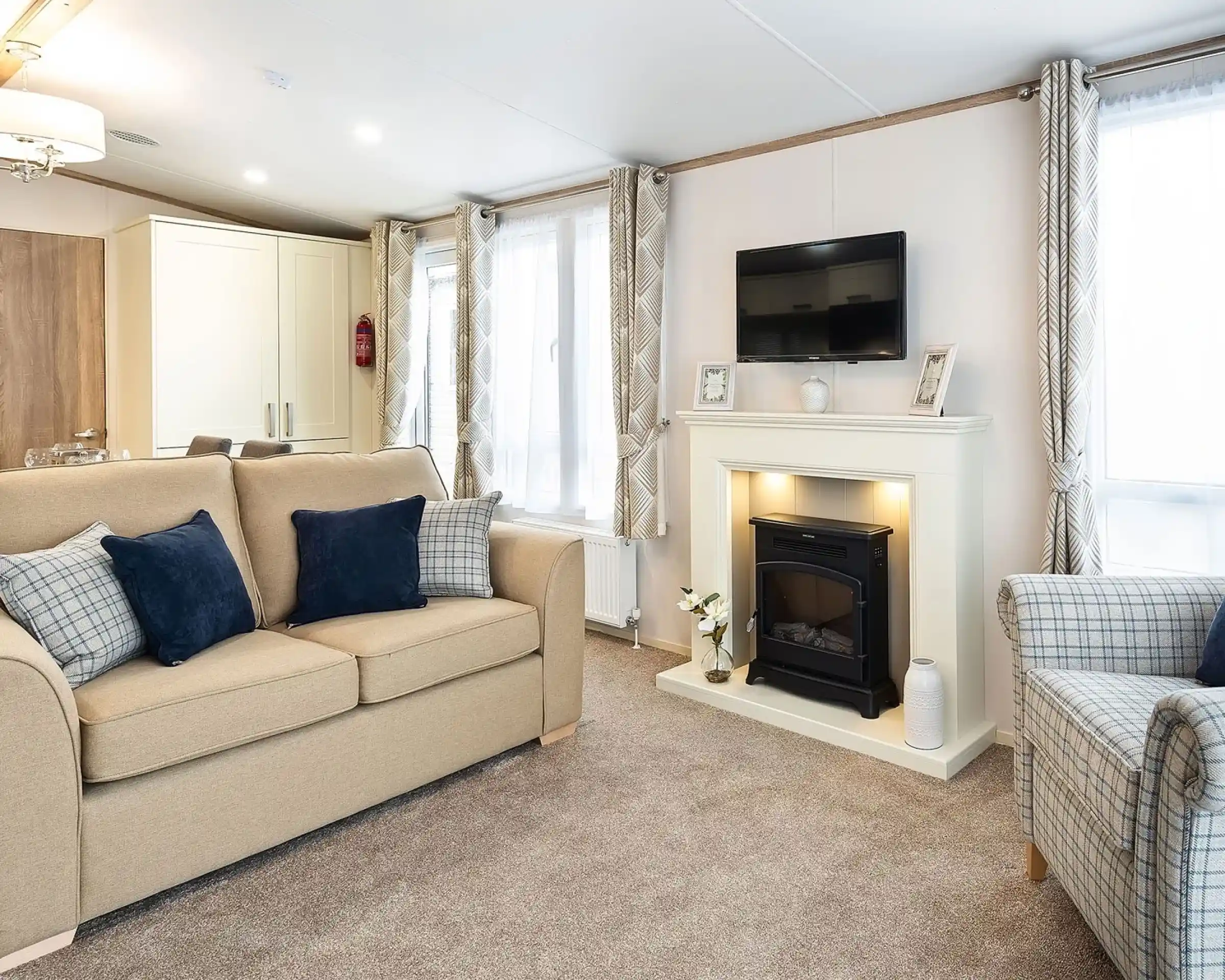 Cozy living room with beige sofa, armchair, and navy and plaid cushions, featuring a white fireplace with built-in electric fire, wall-mounted TV above, and large windows with patterned curtains.
