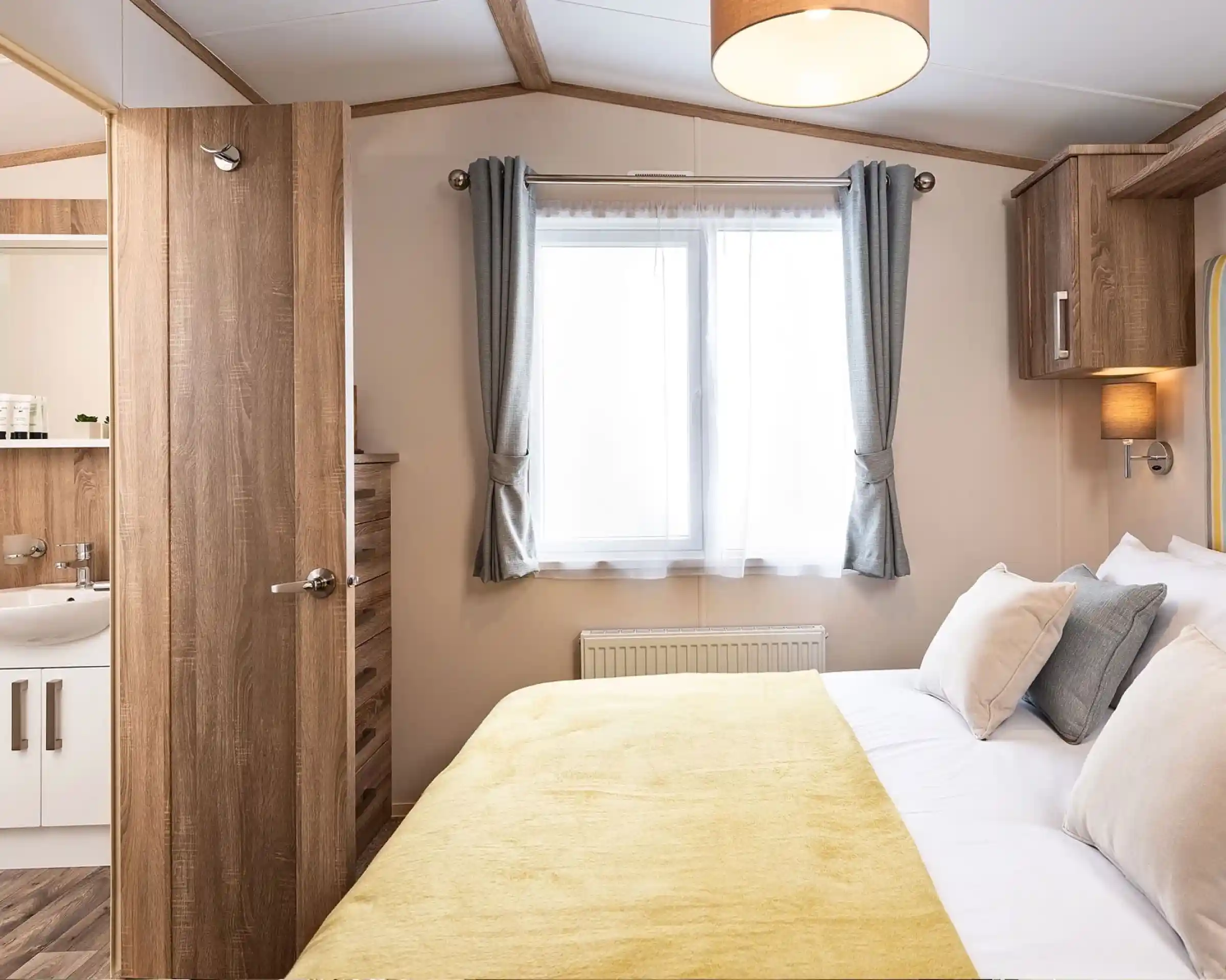 Bright master bedroom featuring a double bed with a yellow throw and white bedding, complemented by beige and gray cushions. A window with sheer white curtains and gray drapes allows natural light into the room. To the left, a wooden door is open, revealing an en-suite bathroom with a white sink and wooden accents. A wooden chest of drawers and overhead storage units add functional charm to the space.