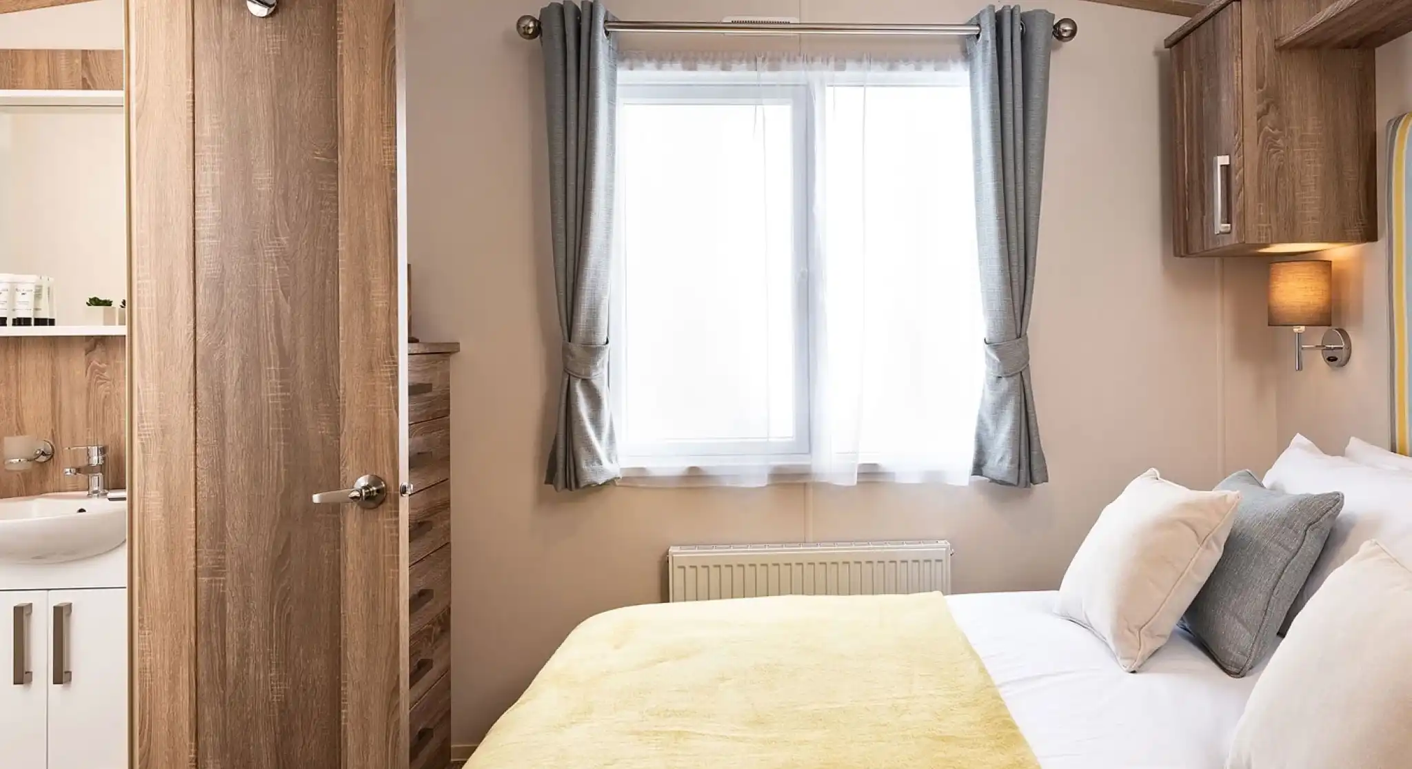 Bright master bedroom featuring a double bed with a yellow throw and white bedding, complemented by beige and gray cushions. A window with sheer white curtains and gray drapes allows natural light into the room. To the left, a wooden door is open, revealing an en-suite bathroom with a white sink and wooden accents. A wooden chest of drawers and overhead storage units add functional charm to the space.