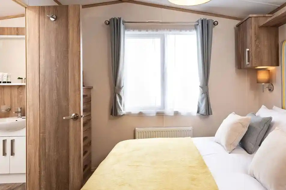 Bright master bedroom featuring a double bed with a yellow throw and white bedding, complemented by beige and gray cushions. A window with sheer white curtains and gray drapes allows natural light into the room. To the left, a wooden door is open, revealing an en-suite bathroom with a white sink and wooden accents. A wooden chest of drawers and overhead storage units add functional charm to the space.