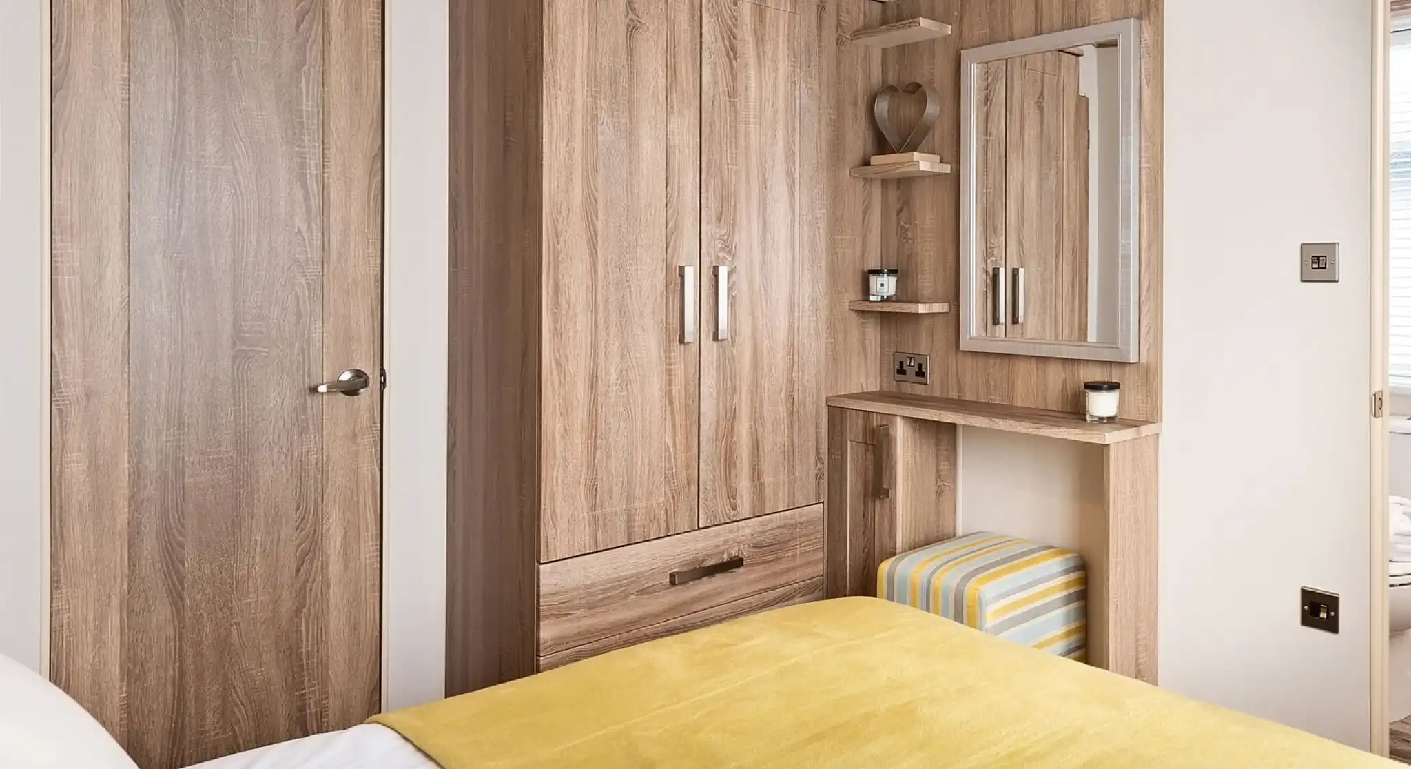Master bedroom featuring a cozy bed with a yellow throw blanket and white sheets. The room includes a built-in wooden wardrobe, a small vanity or dressing table with a mirror, and a striped cushioned stool tucked underneath. Two small floating shelves beside the mirror hold decorative items. A wooden door and a glimpse into the adjoining bathroom are visible.