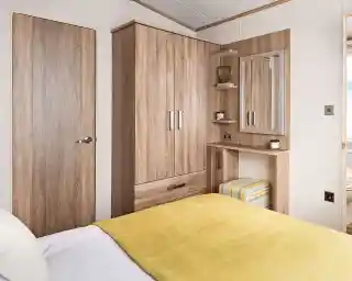 Master bedroom featuring a cozy bed with a yellow throw blanket and white sheets. The room includes a built-in wooden wardrobe, a small vanity or dressing table with a mirror, and a striped cushioned stool tucked underneath. Two small floating shelves beside the mirror hold decorative items. A wooden door and a glimpse into the adjoining bathroom are visible.