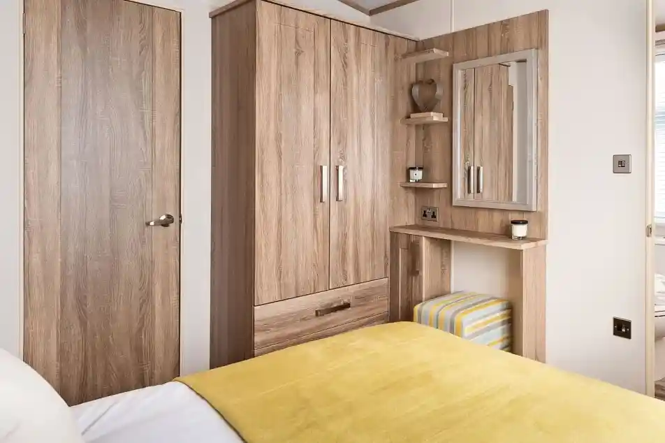 Master bedroom featuring a cozy bed with a yellow throw blanket and white sheets. The room includes a built-in wooden wardrobe, a small vanity or dressing table with a mirror, and a striped cushioned stool tucked underneath. Two small floating shelves beside the mirror hold decorative items. A wooden door and a glimpse into the adjoining bathroom are visible.