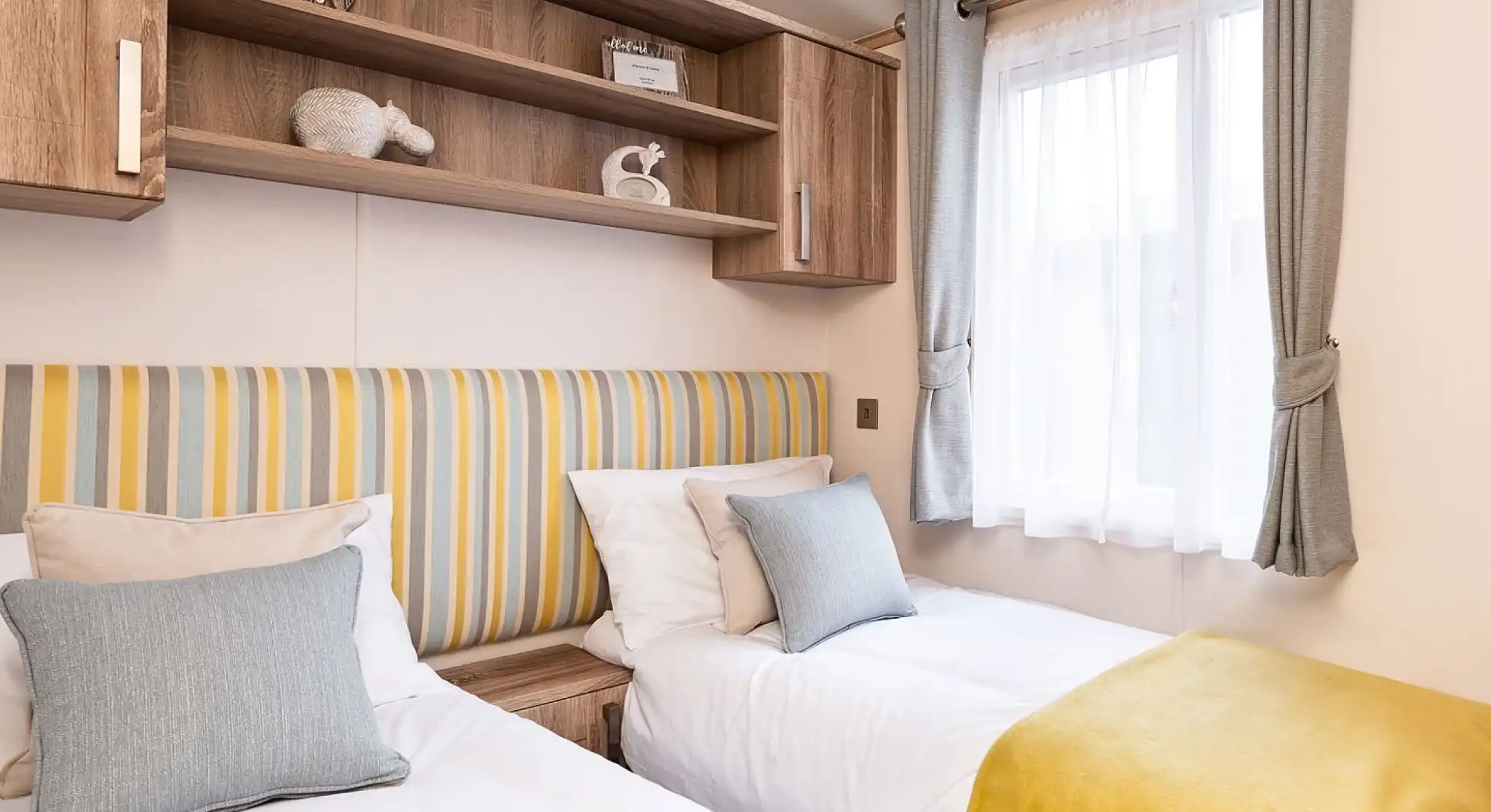 Bright and cozy twin bedroom with two single beds featuring white bedding, yellow throws, and striped yellow and gray headboards. Each bed has a gray and beige cushion. A window with sheer white curtains and gray drapes lets in natural light. Above the beds, there are wooden cabinets and open shelving with decorative items and ornaments.