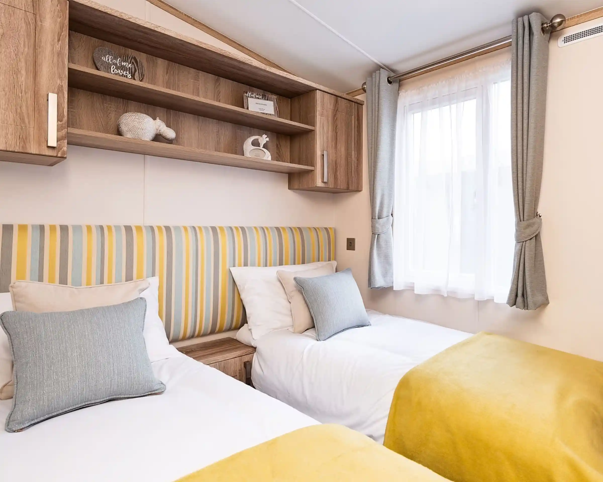 Bright and cozy twin bedroom with two single beds featuring white bedding, yellow throws, and striped yellow and gray headboards. Each bed has a gray and beige cushion. A window with sheer white curtains and gray drapes lets in natural light. Above the beds, there are wooden cabinets and open shelving with decorative items and ornaments.