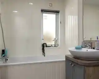 Modern bathroom with a white tub and frosted window. A leafy plant hangs by the window. Sleek sink on a wooden vanity, giving a clean, tranquil feel.