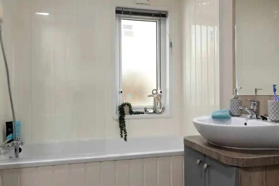 Modern bathroom with a white tub and frosted window. A leafy plant hangs by the window. Sleek sink on a wooden vanity, giving a clean, tranquil feel.