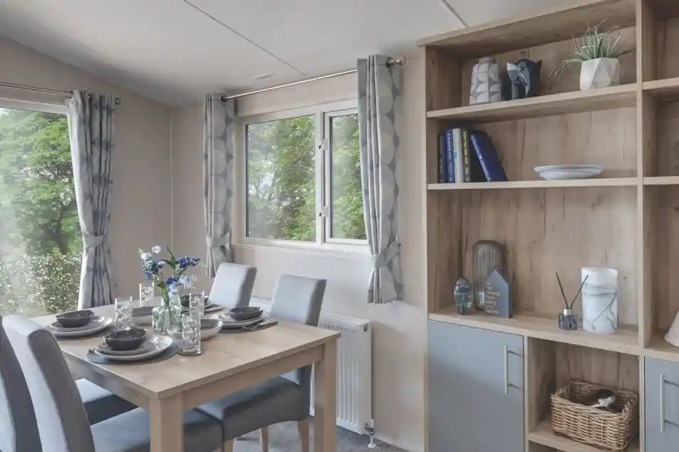 Bright dining area with a wooden table set for four, including grey chairs, plates, glasses, and a flower centerpiece. A wooden shelving unit beside the table displays decorative items, books, and a small plant. The room has light grey curtains and a window with a view of greenery outside.