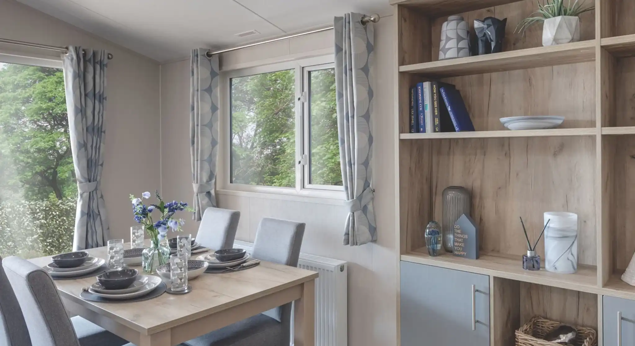Bright dining area with a wooden table set for four, including grey chairs, plates, glasses, and a flower centerpiece. A wooden shelving unit beside the table displays decorative items, books, and a small plant. The room has light grey curtains and a window with a view of greenery outside.