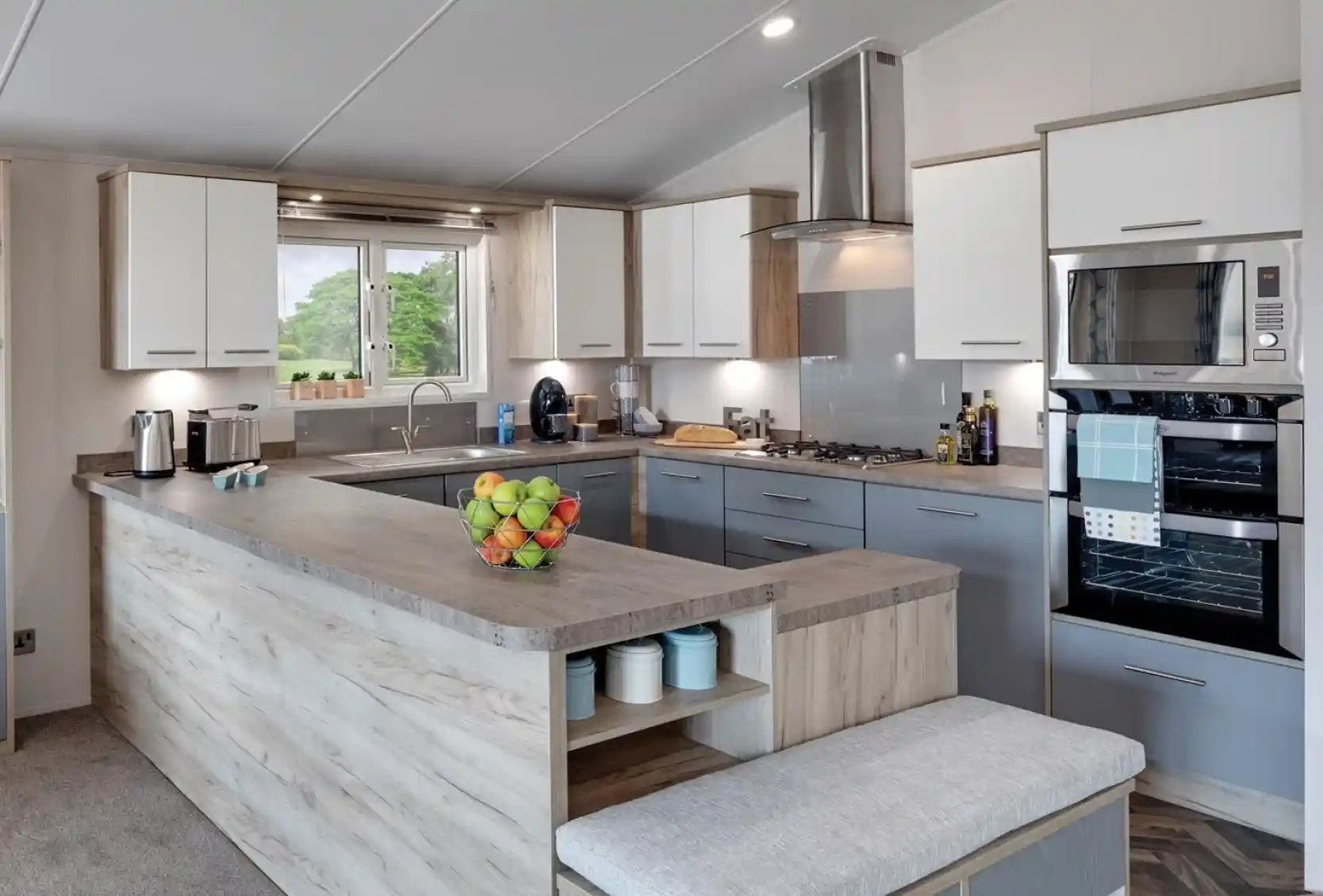 Modern kitchen with a large grey-and-wood island, white and grey cabinetry, stainless steel appliances including a microwave and oven, and a large window offering a view of the outdoors. A bowl of fresh fruit is placed on the countertop, and kitchen essentials like coffee makers and olive oil bottles are visible.