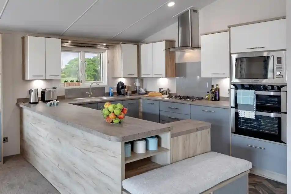 Modern kitchen with a large grey-and-wood island, white and grey cabinetry, stainless steel appliances including a microwave and oven, and a large window offering a view of the outdoors. A bowl of fresh fruit is placed on the countertop, and kitchen essentials like coffee makers and olive oil bottles are visible.