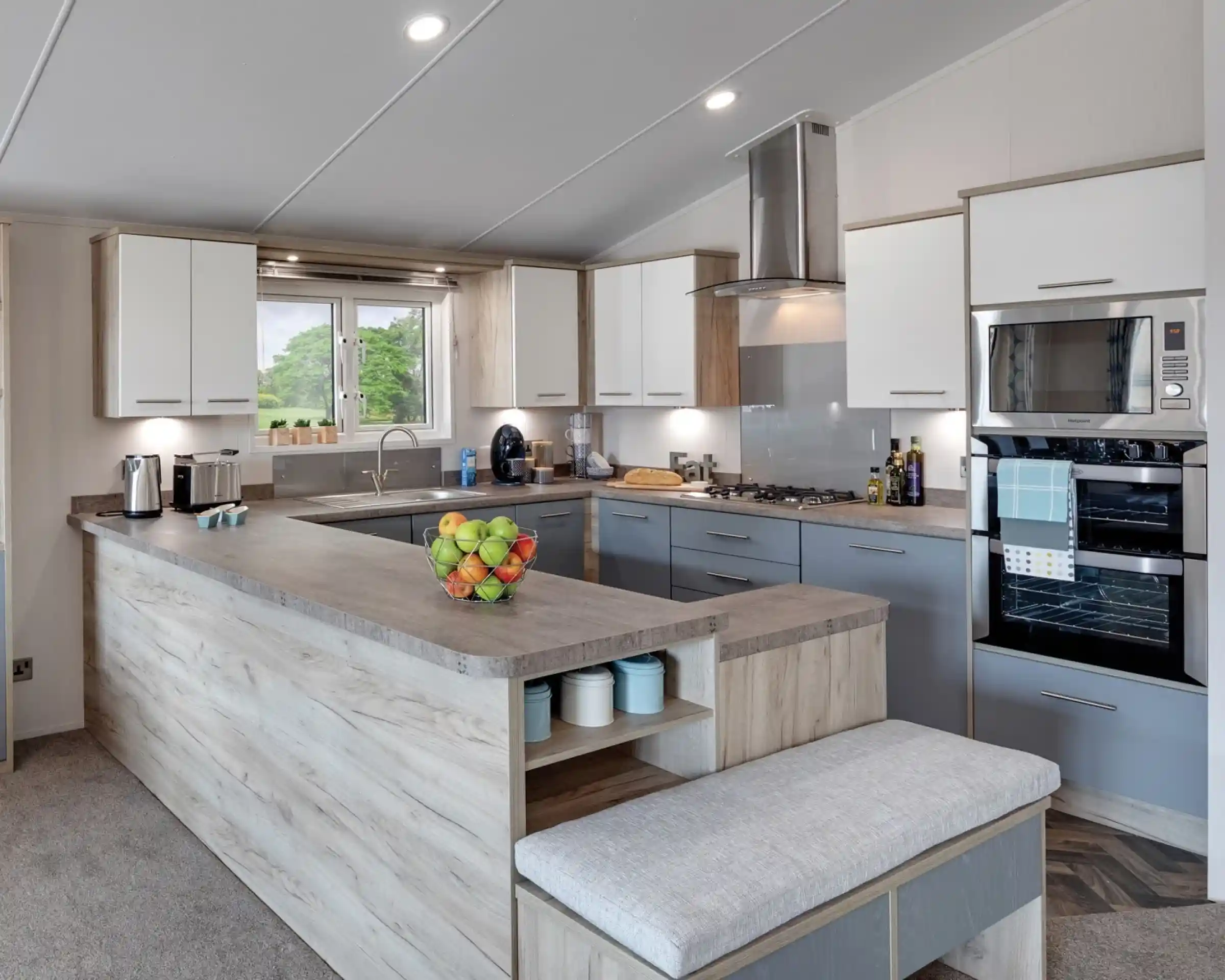 Modern kitchen with a large grey-and-wood island, white and grey cabinetry, stainless steel appliances including a microwave and oven, and a large window offering a view of the outdoors. A bowl of fresh fruit is placed on the countertop, and kitchen essentials like coffee makers and olive oil bottles are visible.