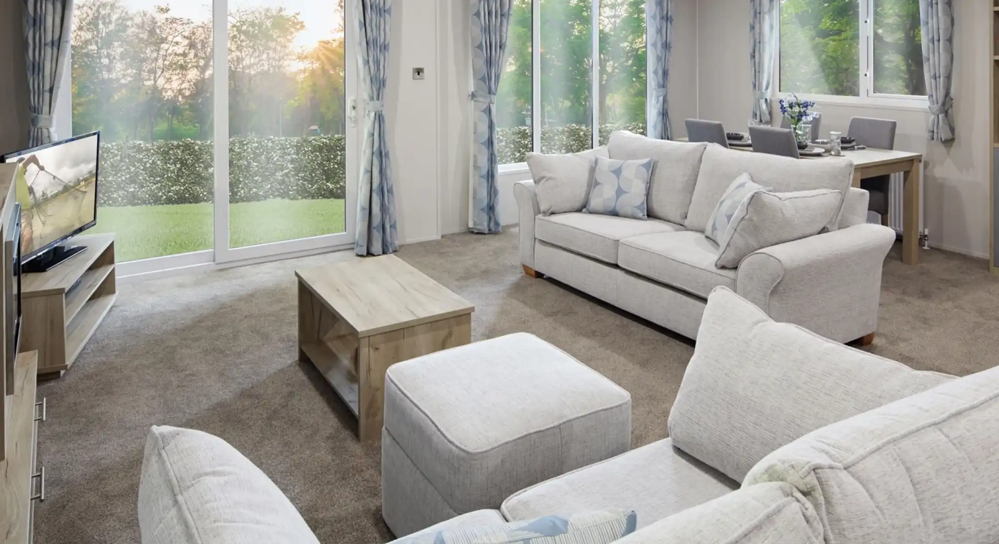 Modern living room with light grey sofas, matching curtains, a flat-screen TV, wooden coffee table, and dining area in the background. Large sliding glass doors and windows let in natural light and offer a view of a green outdoor landscape at sunset.