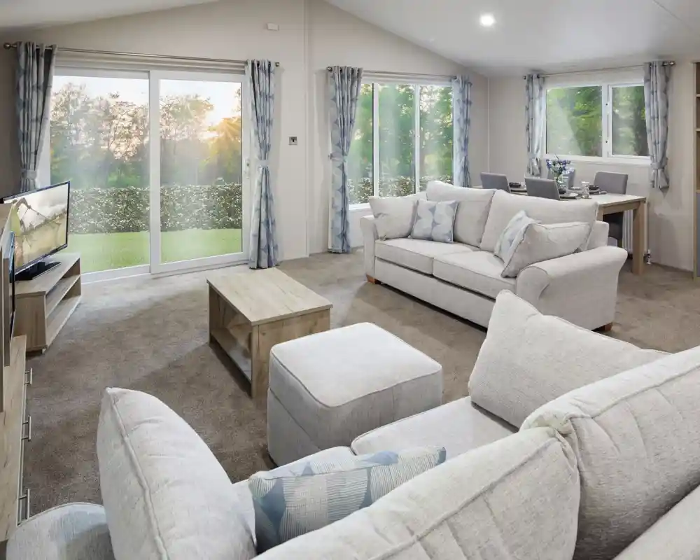 Modern living room with light grey sofas, matching curtains, a flat-screen TV, wooden coffee table, and dining area in the background. Large sliding glass doors and windows let in natural light and offer a view of a green outdoor landscape at sunset.